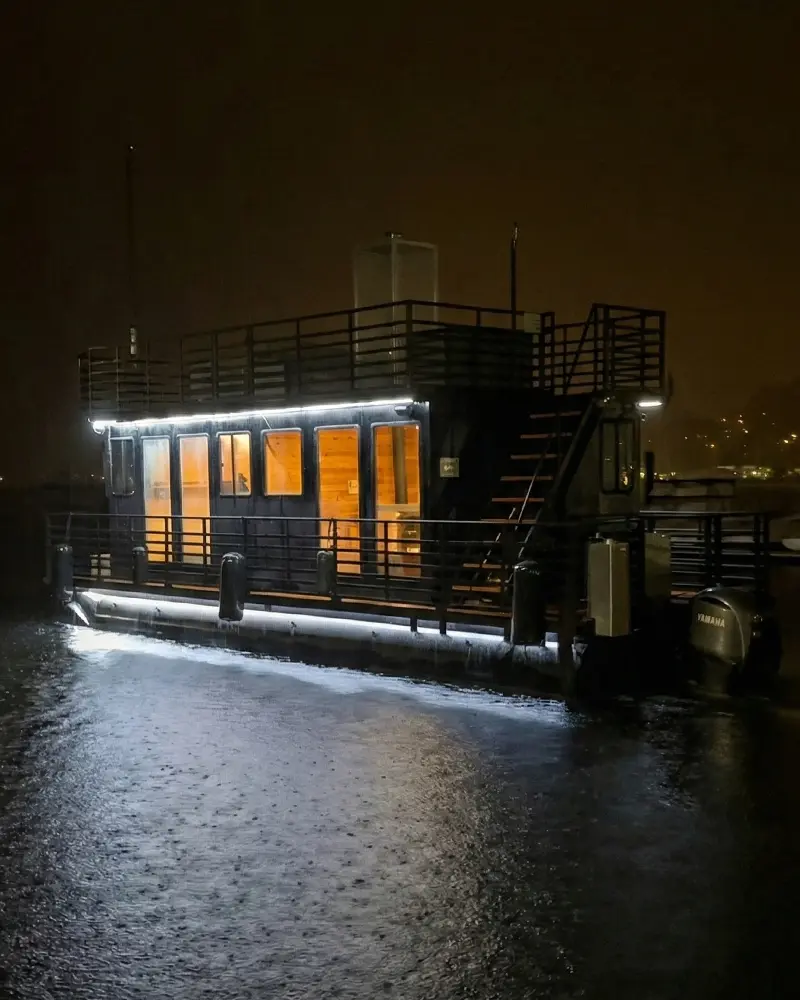 Exterior view of a floating sauna boat at night, with warm cabin lights reflecting on the rainy ocean water.