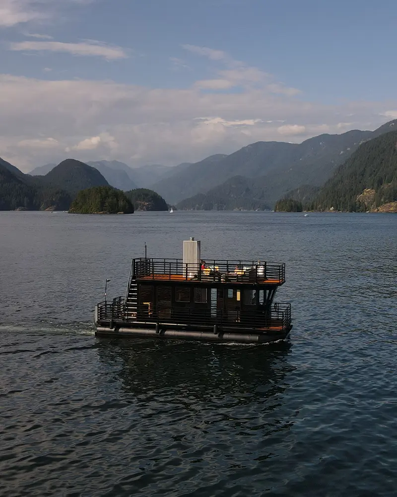 Aerial view of the Hot Voyage floating sauna boat cruising through the deep waters of Indian Arm fjord surrounded by mountains near Vancouver.